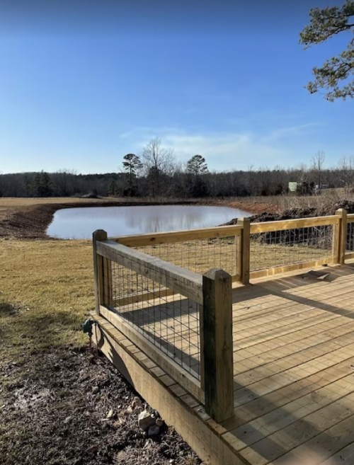 A wooden deck overlooking a calm pond surrounded by trees and open fields under a clear blue sky.