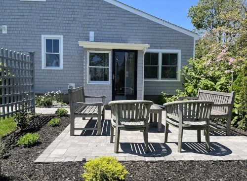 A cozy outdoor seating area with wooden chairs and a stone patio, surrounded by greenery and flowers.