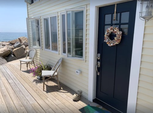 A coastal home entrance with a dark door, seashell wreath, and chairs beside a wooden walkway and rocky shoreline.
