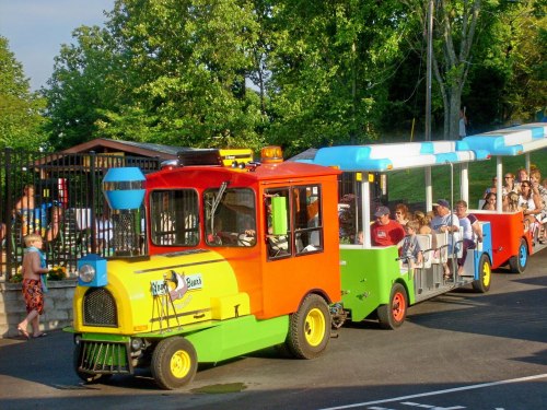 A colorful train-like vehicle with passengers, parked near a pool area surrounded by trees.