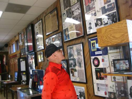 An elderly man in a red shirt and cap gazes at framed photos and memorabilia on a wooden wall in a museum.