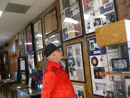 An elderly man in a red shirt and cap gazes at framed photos and memorabilia on a wooden wall in a museum.