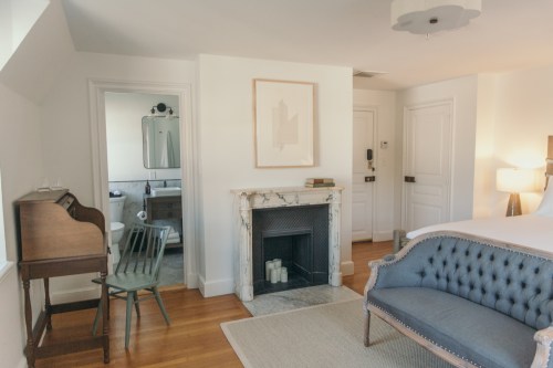 A cozy bedroom featuring a blue sofa, marble fireplace, and a desk, with a bathroom visible in the background.