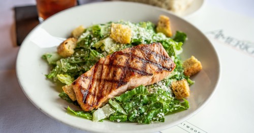 Grilled salmon fillet on a bed of romaine lettuce, croutons, and Parmesan cheese, served in a white bowl.