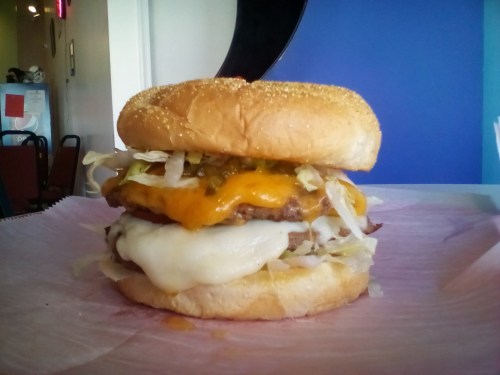 A stacked burger with lettuce, two cheese layers, and a sesame seed bun, resting on pink paper.