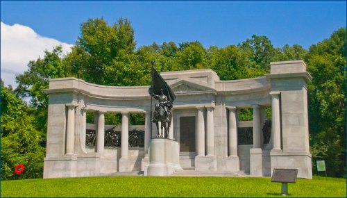 A stone monument with columns and a statue of a soldier holding a flag, surrounded by trees and grass.