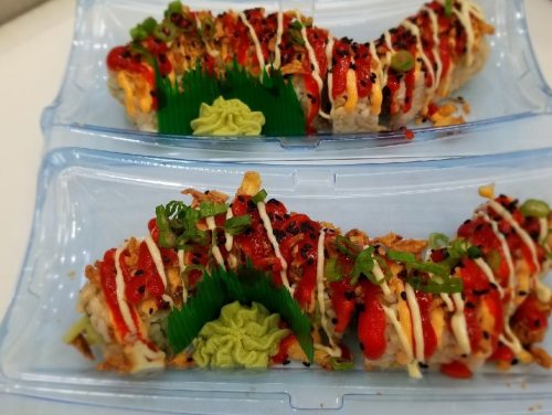 Two sushi rolls topped with sauces, green onions, and wasabi, presented in clear plastic containers.