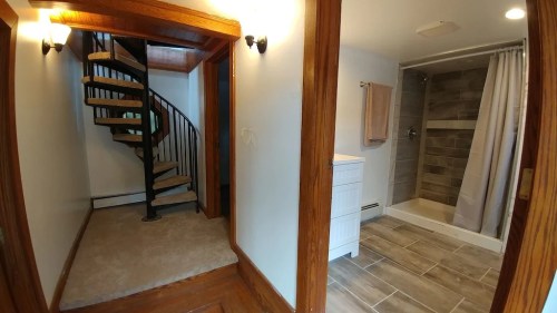 A view of a hallway leading to a bathroom with a shower, featuring a spiral staircase in the background.