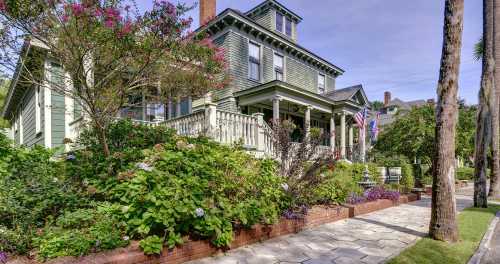 A charming green Victorian house with a porch, surrounded by colorful flowers and trees, on a sunny day.