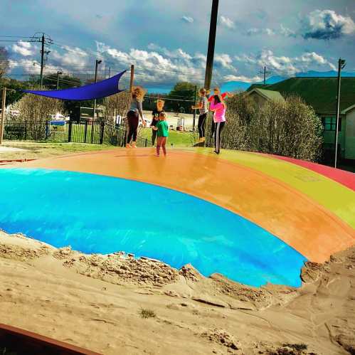 Children play on a colorful inflatable bounce pad at a park under a bright sky with scattered clouds.