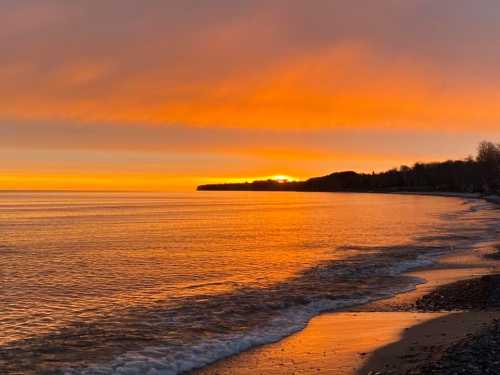 A serene beach at sunset, with vibrant orange and yellow hues reflecting on calm water and a distant shoreline.