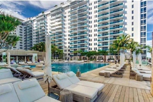 A modern pool area with lounge chairs, surrounded by palm trees and a high-rise building under a clear blue sky.