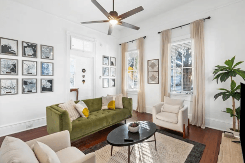 A bright living room with a green sofa, white armchair, artwork on the walls, and large windows letting in natural light.
