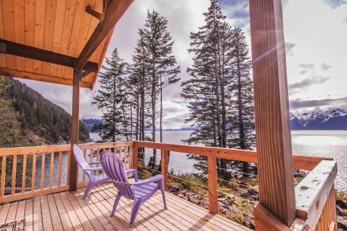 A scenic view from a wooden deck with two purple chairs, overlooking a lake surrounded by trees and mountains.