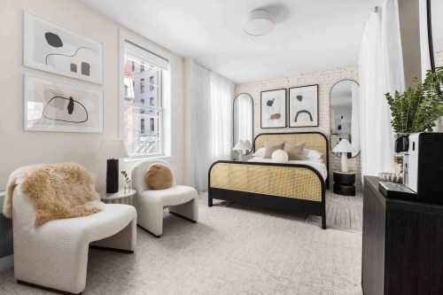 Cozy bedroom with a bed, two chairs, artwork on the walls, and large windows letting in natural light.