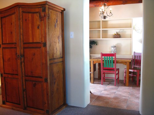 A wooden armoire stands beside a dining area with a table and colorful chairs in a cozy, well-lit room.