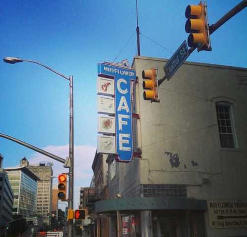 A vintage café sign reading "Mayflower Café" at a street corner with traffic lights and a clear blue sky.