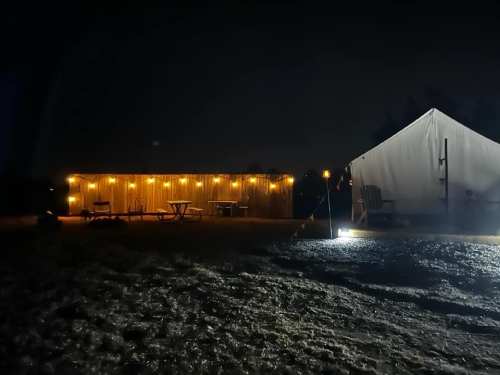 A dimly lit campsite at night featuring a cabin with string lights and a large tent nearby.