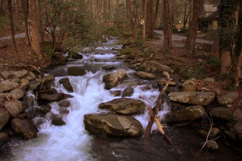 A serene stream flows over rocks, surrounded by trees, with a glimpse of a cabin in the background.