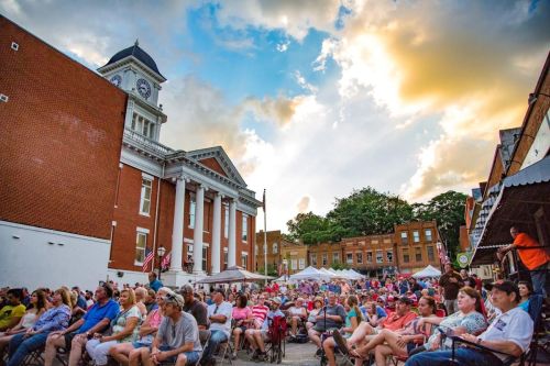 A large crowd gathers in front of a historic building during an outdoor event, with a colorful sky above.