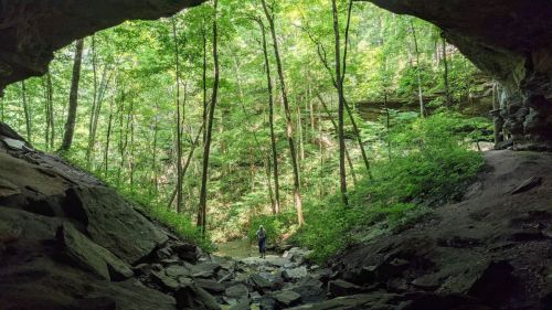 A person stands at the entrance of a cave, surrounded by lush green trees and rocky terrain.