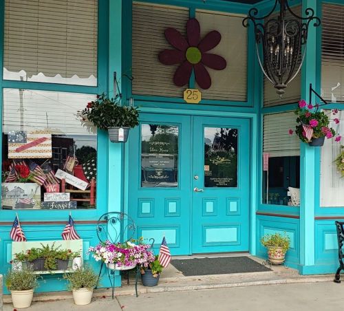 Brightly colored storefront with a large flower decoration, potted plants, and American flags adorning the entrance.