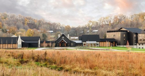 A modern building complex surrounded by autumn foliage and grassy fields, set against a hilly landscape.