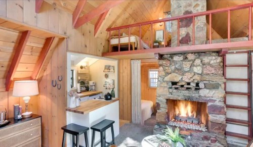 Cozy cabin interior with a stone fireplace, wooden beams, and a lofted sleeping area. Warm and inviting atmosphere.