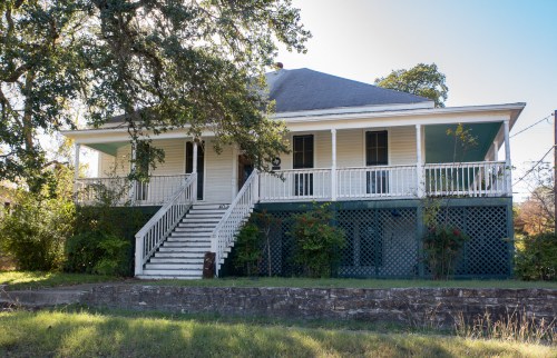 A historic two-story house with a porch, white siding, and a staircase, surrounded by greenery and a stone wall.