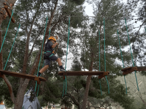 A child in a helmet navigates a rope bridge among trees at a camp, showcasing adventure and outdoor activity.