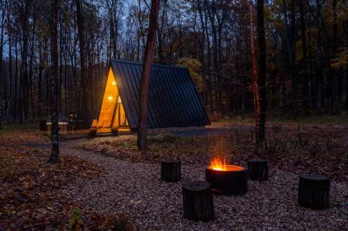A modern cabin with warm light at dusk, surrounded by trees and a fire pit with glowing flames in a serene forest setting.