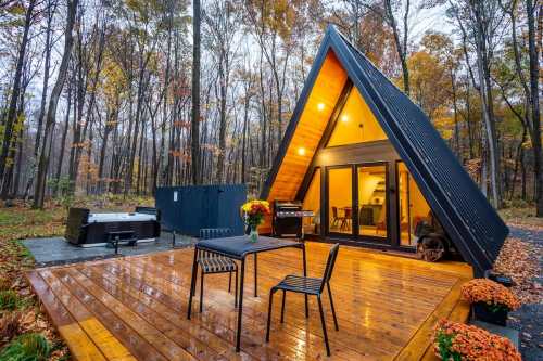 A modern A-frame cabin in a forest, featuring a wooden deck, outdoor seating, and a hot tub, surrounded by autumn foliage.
