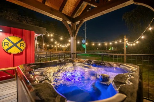 A hot tub with blue lighting under a wooden gazebo, surrounded by string lights and a railroad sign at night.