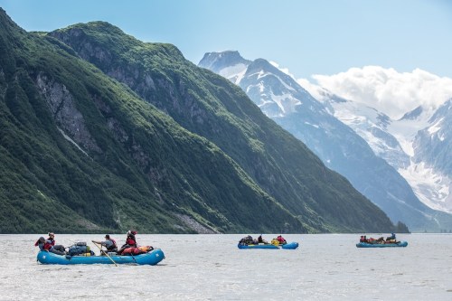 Four rafts float on a river surrounded by lush green mountains and snow-capped peaks under a clear blue sky.