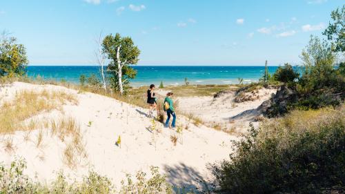 Two people walking on sandy dunes with a view of a blue lake and clear sky in the background.
