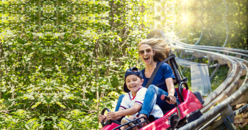 A woman and a child joyfully ride a roller coaster through a lush green forest.