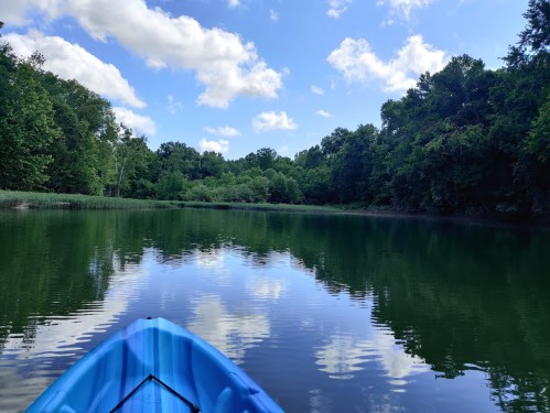 A blue kayak on calm water, reflecting trees and clouds under a bright sky. Lush greenery surrounds the serene scene.