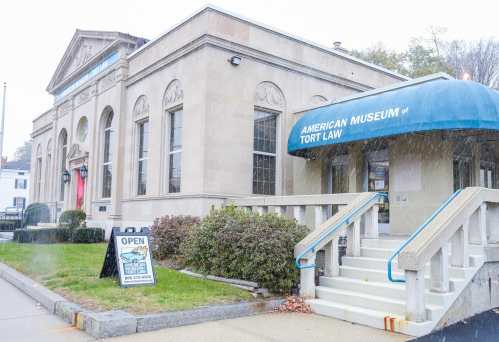 Exterior of the American Museum of Tort Law, featuring a blue awning and a sign indicating it's open, with light snow falling.