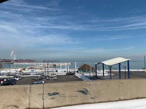 A snowy waterfront scene with a parking lot, a pavilion, and a view of the lake under a clear blue sky.