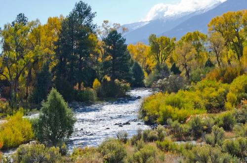 A serene river flows through a vibrant landscape of autumn trees and mountains under a clear blue sky.
