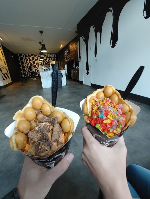 Two hands hold waffle bowls filled with colorful toppings: one with chocolate and the other with fruity cereal.