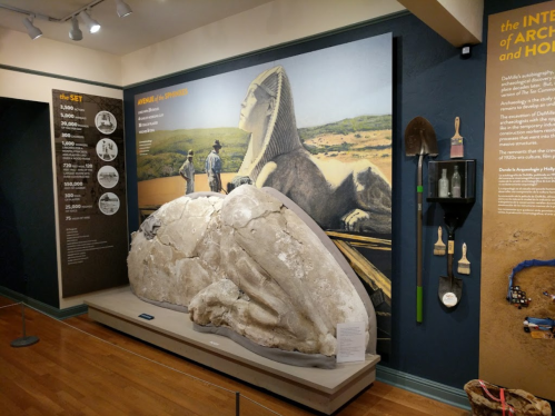 Exhibit featuring a large stone sculpture, tools, and a mural of a sphinx in an archaeological display.