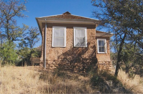 A small, rustic house with a stone foundation, surrounded by dry grass and trees under a clear blue sky.