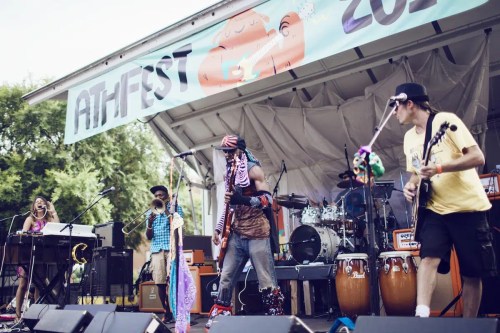 A lively band performs on stage at a festival, with musicians playing various instruments and a colorful backdrop.