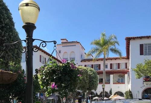 A sunny street scene featuring a lamp post, hanging flower baskets, and a building with palm trees in the background.