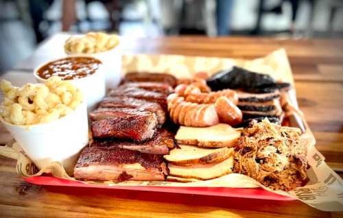 A wooden table with a tray of assorted barbecue meats, sides of macaroni and cheese, and baked beans.