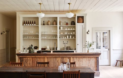 A rustic bar area with a wooden counter, shelves of glassware, and a large dining table in a bright, minimalist space.