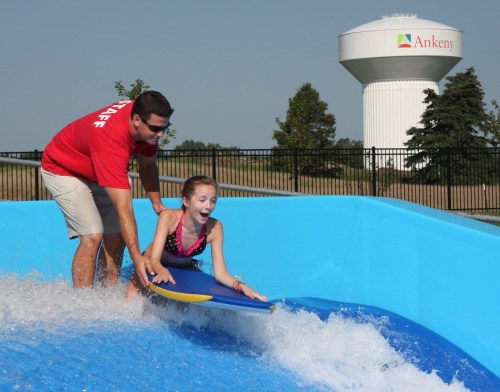 A staff member assists a girl on a surfboard in a wave pool, with a water tower in the background.