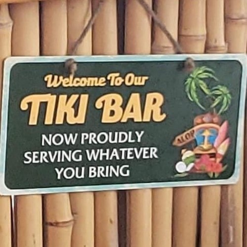 Sign for a tiki bar featuring tropical imagery, stating "Welcome To Our TIKI BAR - NOW PROUDLY SERVING WHATEVER YOU BRING."