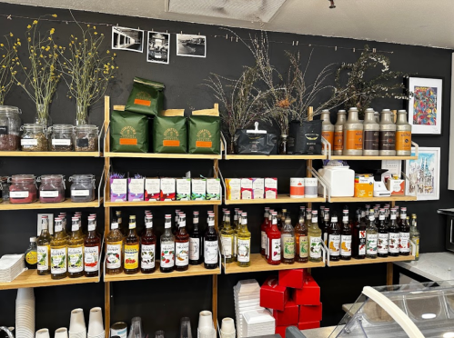 A shelf displaying various jars, bags, and bottles of beverages and ingredients in a cozy shop setting.